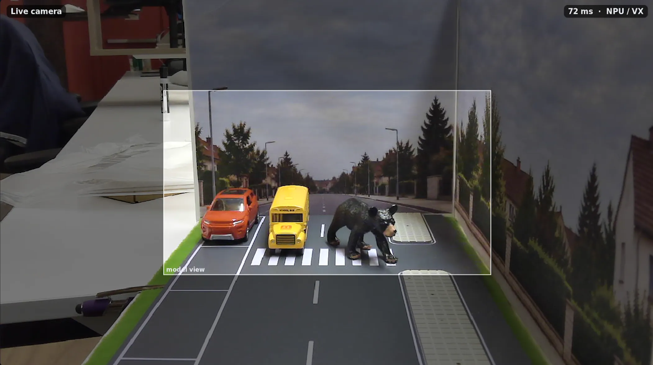 Live camera view of a model road diorama: an orange car, a school bus, and a bear figure crossing at a crosswalk.